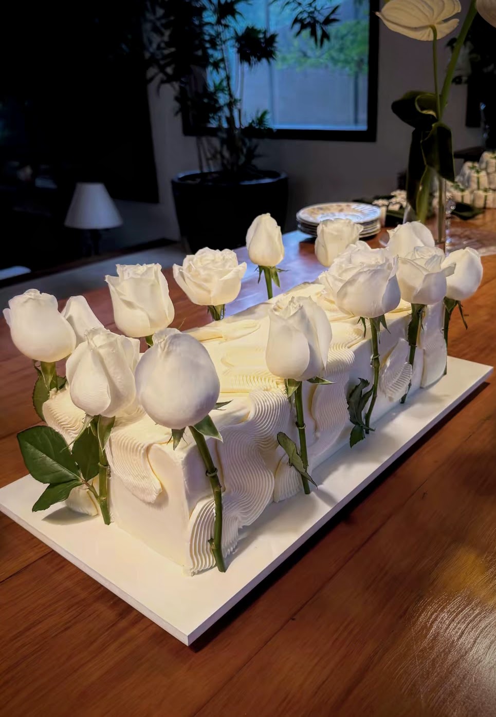 Bolo retangular alto em buttercream branco com textura de ondas e estrias, atravessado por rosas brancas naturais de cabo longo, sobre base branca em mesa de madeira