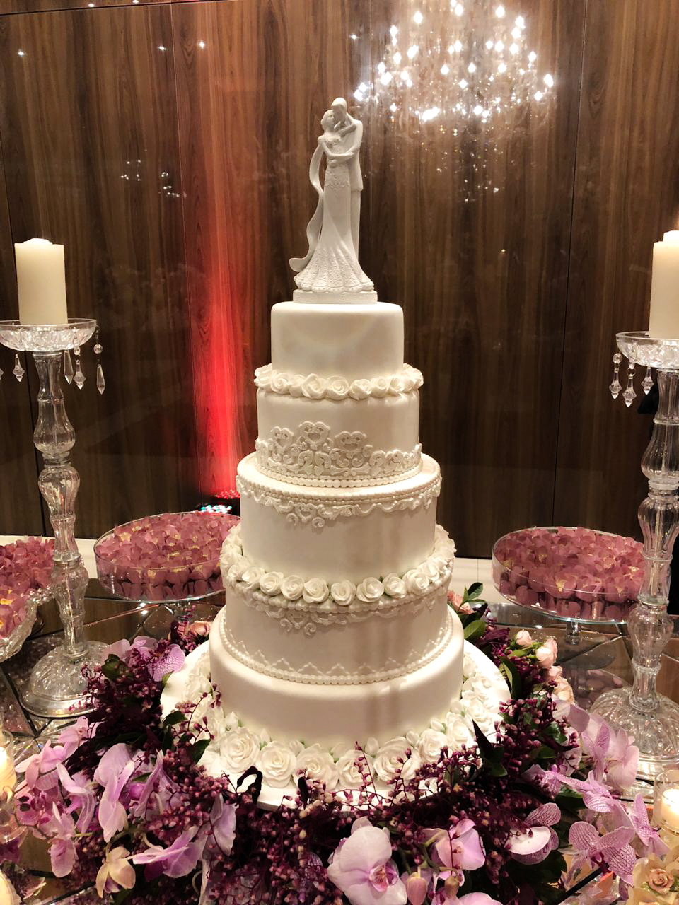 Bolo de casamento branco de cinco andares em pasta americana, com faixas de rosas modeladas, rendas em glacê real e fileiras de pérolas entre os andares, topo de noivos e orquídeas ao redor da base
