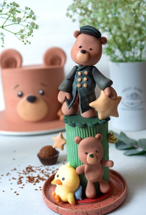 Bolo escultórico com urso de casaco xadrez e boina modelado à mão, sobre base cilíndrica verde, com estrelas e patinho em pasta americana