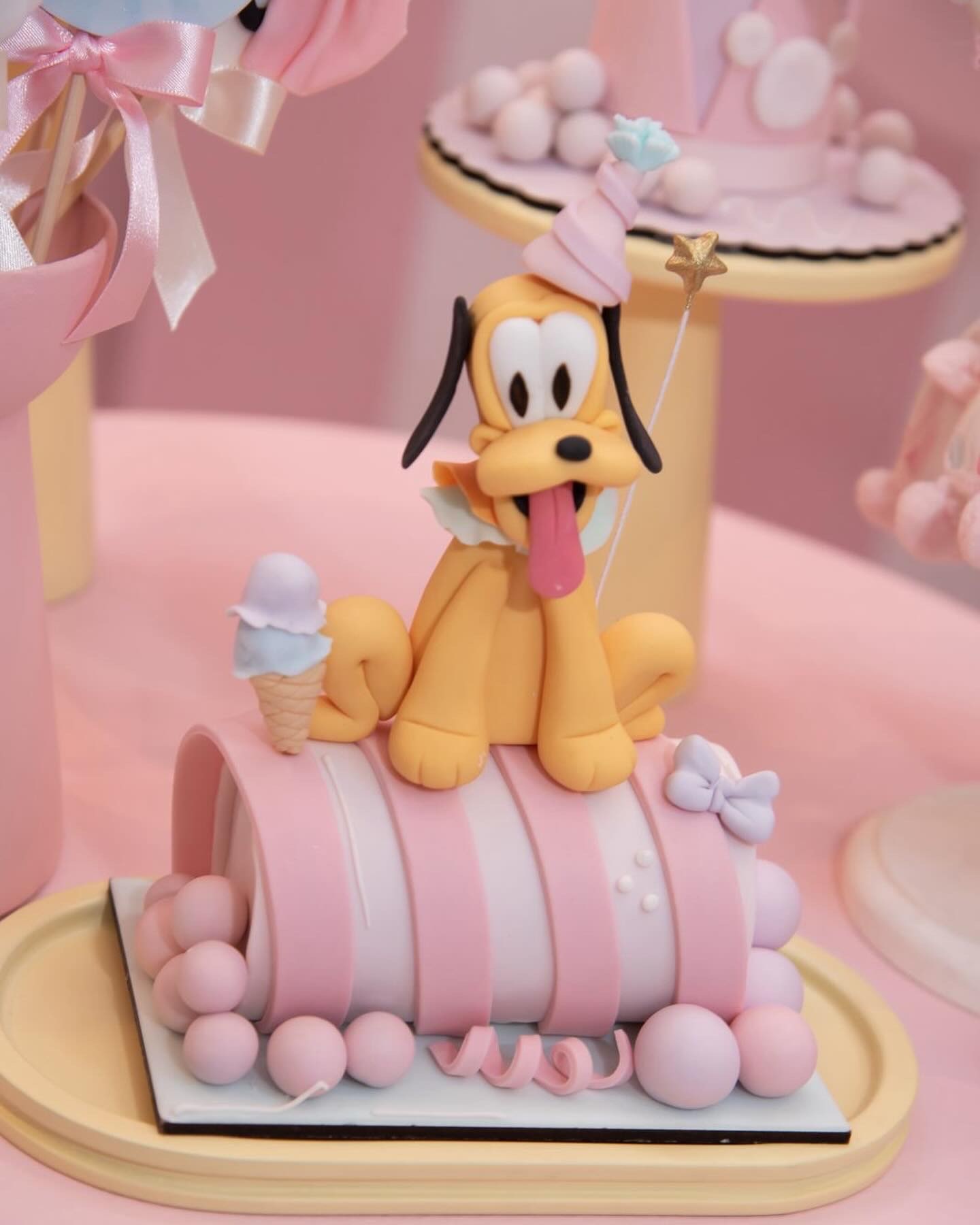 Bolo cilíndrico deitado em listras rosa e branco, com cachorro modelado à mão no topo, casquinha de sorvete e esferas de pasta americana ao redor