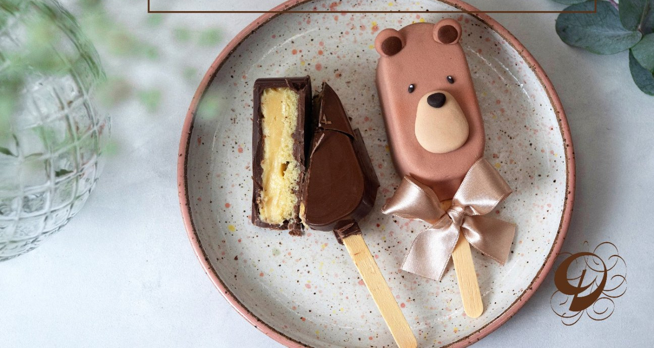 Popsicle de bolo com carinha de urso e laço de cetim ao lado de outro popsicle cortado, mostrando a massa de baunilha com recheio