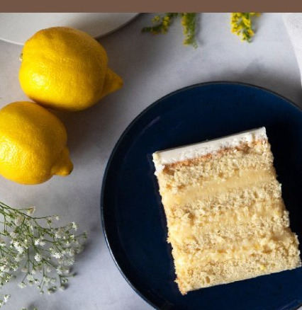 Fatia de bolo de limão-siciliano em prato azul-marinho, com dois limões e flores ao lado